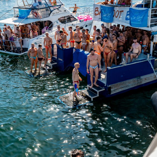 A lively scene at a boat party on the water, hosted by Sydney Harbour Boat Hire The Yacht Social Club. People in swimsuits are on deck and in the water, with several yachts docked together in a picturesque coastal area, city buildings visible in the background.