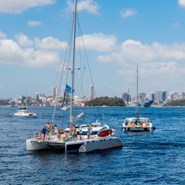 A group of people enjoys a sunny day on a catamaran sailboat named "Dawn Patrol" on a clear blue body of water. Other boats are visible in the background, with a city skyline and partly cloudy sky. The logo "The Yacht Social Club Sydney Boat Hire" is in the bottom right corner.