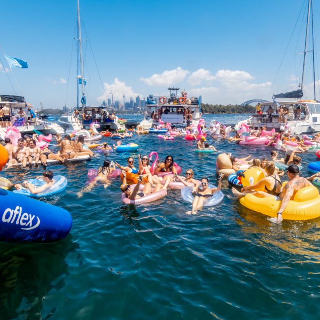 A crowded gathering of people enjoying a sunny day on the water, floating on colorful inflatables near docked boats. Some are in unicorn and donut floaties. The city skyline is visible in the background under a clear blue sky, showcasing the charm of The Yacht Social Club Sydney Boat Hire.