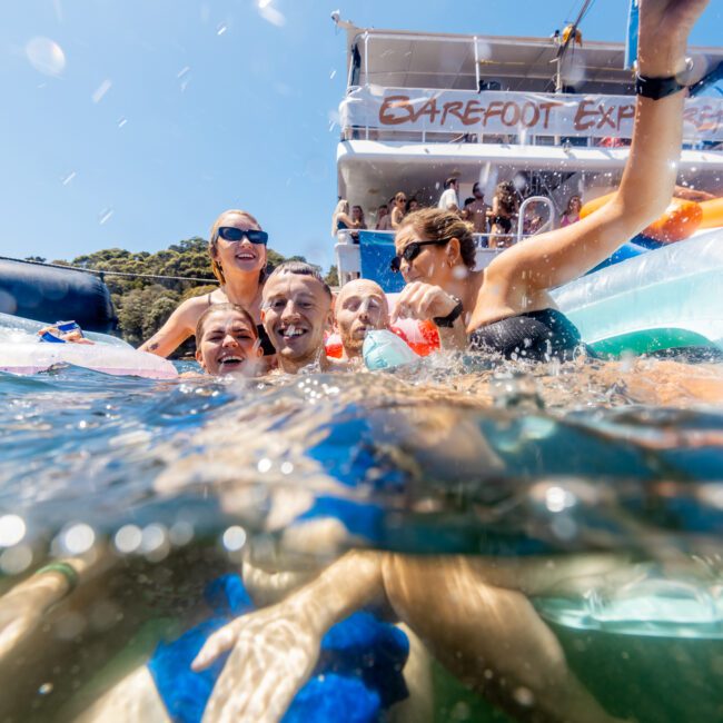 A group of people in swimwear joyfully splashing in the water near a boat. The waterline is just below their shoulders, showing their excited faces. The boat behind them has a sign that reads "BAREFOOT EXP." Another person from The Yacht Social Club Event Boat Charters is diving into the water nearby.