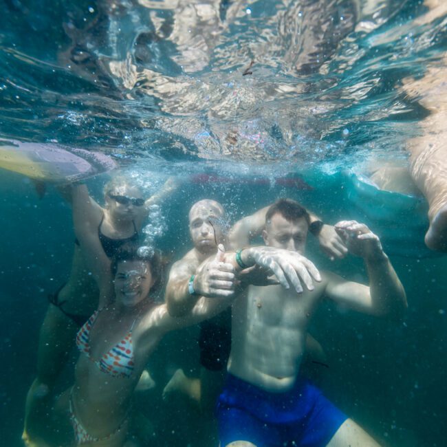 A group of four people are swimming underwater, smiling and posing for the camera. They are in a clear, blue body of water. The image is lively, with reflections and bubbles visible above them. "Luxury Yacht Rentals Sydney" is written in white text on the bottom right.