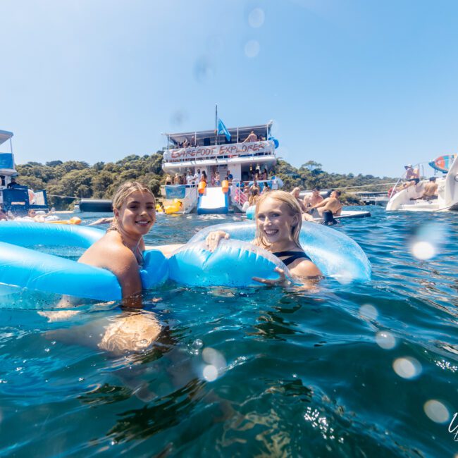 Two women float on inflatable loungers in a clear blue sea, smiling at the camera. Behind them, several people enjoy a sunny day on large yachts and boats. Trees and land are visible in the background. A logo in the corner reads "The Yacht Social Club," promoting Luxury Yacht Rentals Sydney.