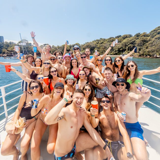 A large group of people in swimsuits is gathered on a boat, smiling and celebrating with drinks in hand. They are surrounded by water and greenery, suggesting a sunny day on a lake or river. The atmosphere is joyful and festive as several people wave at the camera, embodying the vibrant spirit of Boat Parties Sydney The Yacht Social Club.