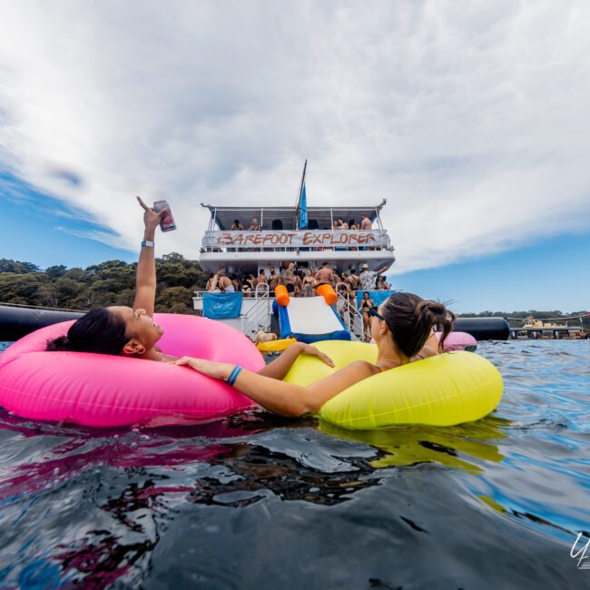 Two people float on pink and yellow inflatable rings in the water, holding hands. They face a large boat party with a banner reading "Barefoot Explorer." The sky is partly cloudy, and attendees are visible on the multi-level boat by The Yacht Social Club Sydney Boat Hire.