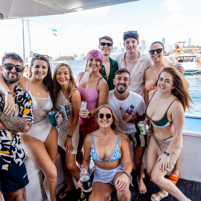 A group of ten people, smiling and posing on a boat on a sunny day. They are dressed in swimwear, enjoying a day on the water with The Yacht Social Club. The background shows other boats and a city skyline under a clear blue sky.