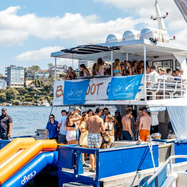 A lively group of people are enjoying a sunny day on a boat party hosted by The Yacht Social Club Sydney Boat Hire. The boat, adorned with banners that read "Afloat," is docked by the waterfront. Some attendees socialize on the lower deck while others relax on the upper deck, with a yellow and blue inflatable slide visible.