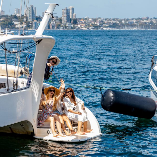 Three women sitting at the back of a white yacht, smiling and waving toward the camera. They're wearing sun hats and sunglasses, with water and cityscape visible in the background. The Yacht Social Club offers unforgettable boat parties in Sydney, perfect for days like this on luxurious yachts.