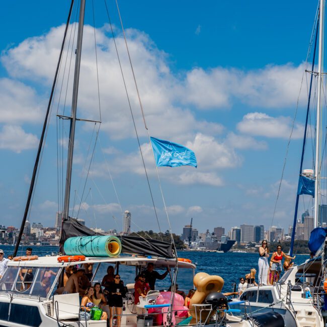 A catamaran filled with people is anchored in a sunny harbor. A blue flag flies at the top of the mast. In the background, Sydney Harbour Boat Hire's city skyline and a suspension bridge stand tall beneath scattered clouds, offering an idyllic scene for those enjoying Luxury Yacht Rentals Sydney.