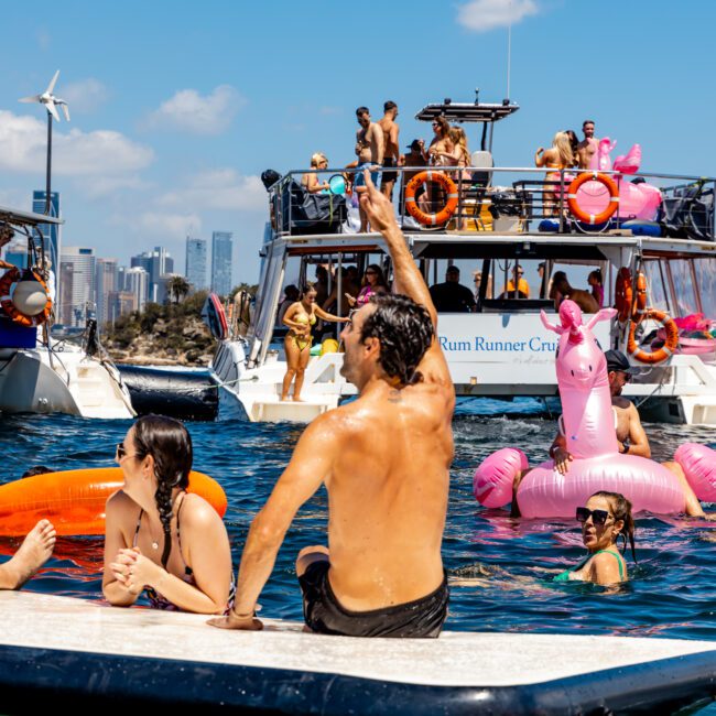 People enjoy a sunny day on the water. A man on a floating platform points upwards while others swim nearby. Behind, a pink flamingo float and a boat with 'Rum Runner Cruise' banner are visible. City buildings and wind turbines stand in the background, making it perfect for The Yacht Social Club Sydney Boat Hire events.