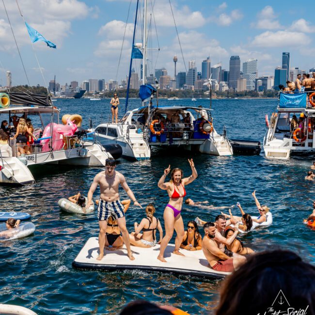 A lively summer party with people having fun on a floating platform and inflatables in the water beside docked boats. The city skyline is visible under a partly cloudy sky, enhancing the festive atmosphere. "The Yacht Social Club Sydney Boat Hire" logo is visible in the corner.