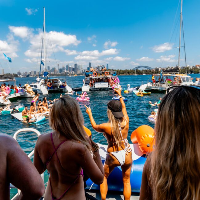 A lively boat party on a sunny day with people in swimwear enjoying the water, hosted by The Yacht Social Club Sydney Boat Hire. Boats and inflatables are scattered around, and the city skyline is visible in the background. A person in the foreground raises their arms, contributing to the festive atmosphere.