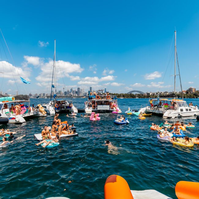 A vibrant scene of The Yacht Social Club is depicted. Numerous people are swimming or lounging on floating devices in the water. Several boats and yachts are gathered around, with the city skyline visible in the background under a clear blue sky.