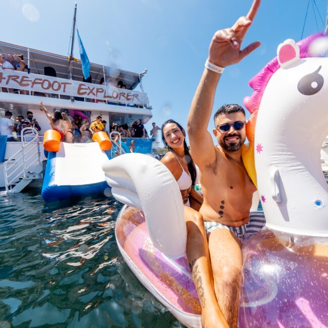 A man and woman smile and pose on an inflatable unicorn float in the water with a boat named "Barefoot Explorer" in the background. The man has his arm raised, and the woman is sitting behind him. The sun shines brightly in the clear blue sky, perfect for Sydney Harbour Boat Hire with The Yacht Social Club.