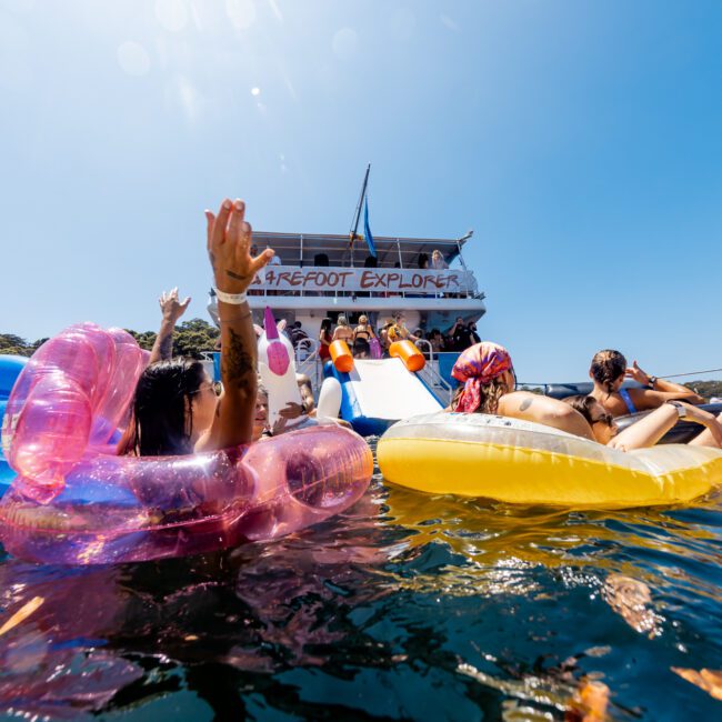 People relax and enjoy the water with inflatable tubes and floats near a large boat labeled "Barefoot Explorer." The clear blue sky and lush greenery form the backdrop of the vibrant social scene. The image captures the essence of a fun-filled day on the water, exemplifying Boat Parties Sydney The Yacht Social Club.