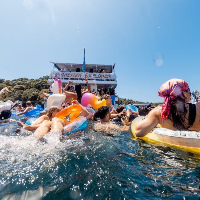 People swimming and floating on inflatables in the sea near a boat named "Barefoot Explorer". Many are wearing swimwear, and the atmosphere is lively and sunny. Additional boats from The Yacht Social Club Event Boat Charters are visible in the background, with the shoreline and trees in the distance.