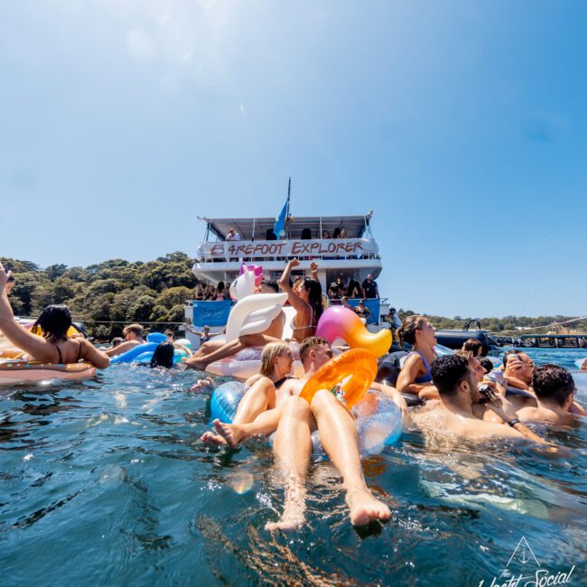 A lively scene with people enjoying a party on inflatable floats in the water near a boat named "Barefoot Explorer." The boat has two visible decks filled with more partygoers. The backdrop features a tree-covered coastline under a clear blue sky, capturing the vibrant essence of The Yacht Social Club Sydney Boat Hire event.