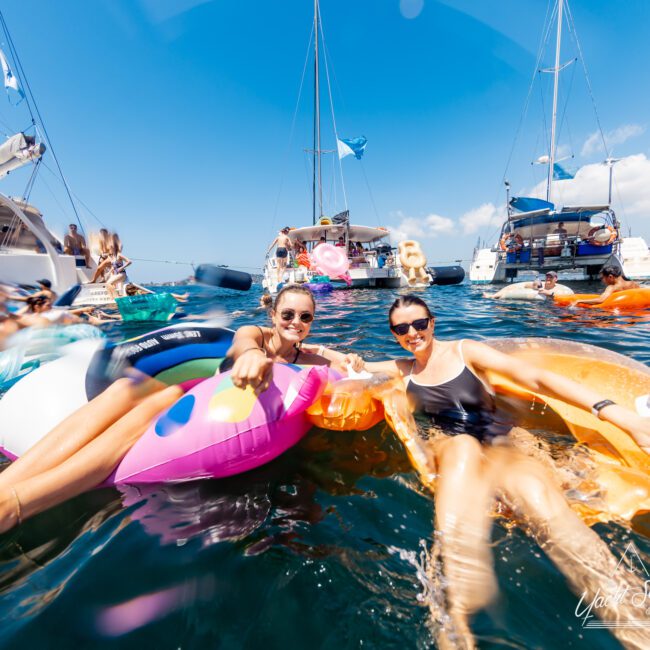 Two women float on colorful inflatable loungers in clear blue water, smiling towards the camera. Behind them, several boats from The Yacht Social Club are anchored with people relaxing. The sky is clear and sunny, creating a vibrant and festive atmosphere perfect for boat parties in Sydney Harbour.