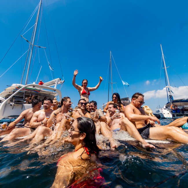 A large group enjoys a sunny day on the water, some in the sea and some on yachts. They are smiling, laughing, and holding drinks. Yachts dot the background under blue skies with a few clouds overhead—perfect for a Sydney Harbour Boat Hire experience with The Yacht Social Club.