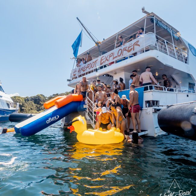 A group of people gather on the back deck of a yacht named "Barefoot Explorer." Some are sitting on an inflatable slide and a large inflatable yellow duck, while others are laughing and having fun in the water. The sun shines brightly, indicating a beautiful day at one of the Boat Parties Sydney The Yacht Social Club organizes.