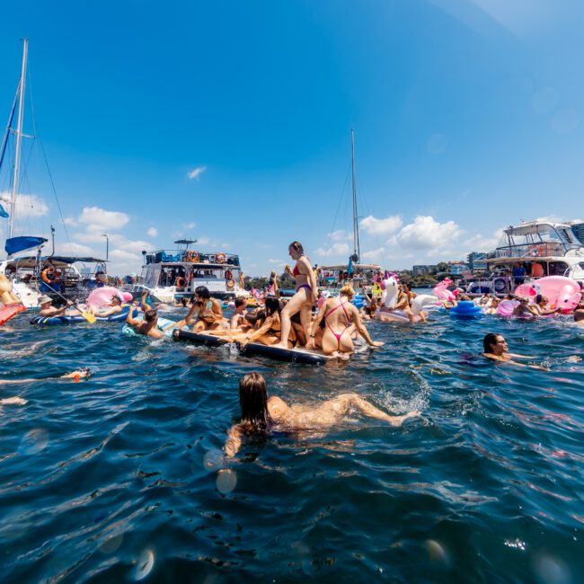 A large group of people enjoying a sunny day on the water, surrounded by boats and colorful inflatable floats. Many individuals are swimming and relaxing. The sky is clear with a few clouds, and the water is filled with vibrant activity and joy during The Yacht Social Club Event Boat Charters.