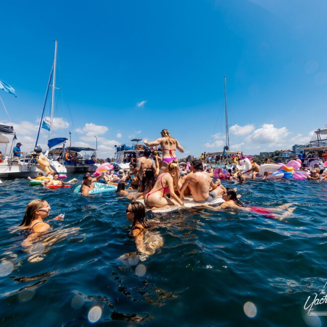 People are enjoying a sunny day on the water, surrounded by boats and various flotation devices. Some are in swimsuits, lounging and socializing. The atmosphere is festive with clear blue skies and calm waters. Balloons and flags are visible in the background at The Yacht Social Club Sydney Boat Hire event.