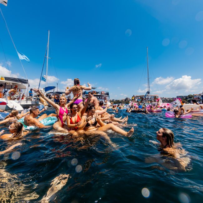 A lively scene of people partying and swimming in the water between multiple yachts. Some are floating on inflatables, while others socialize and splash around. The sun is shining brightly, and the sky is clear with scattered clouds. A logo in the corner reads "The Yacht Social Club Sydney Boat Hire.