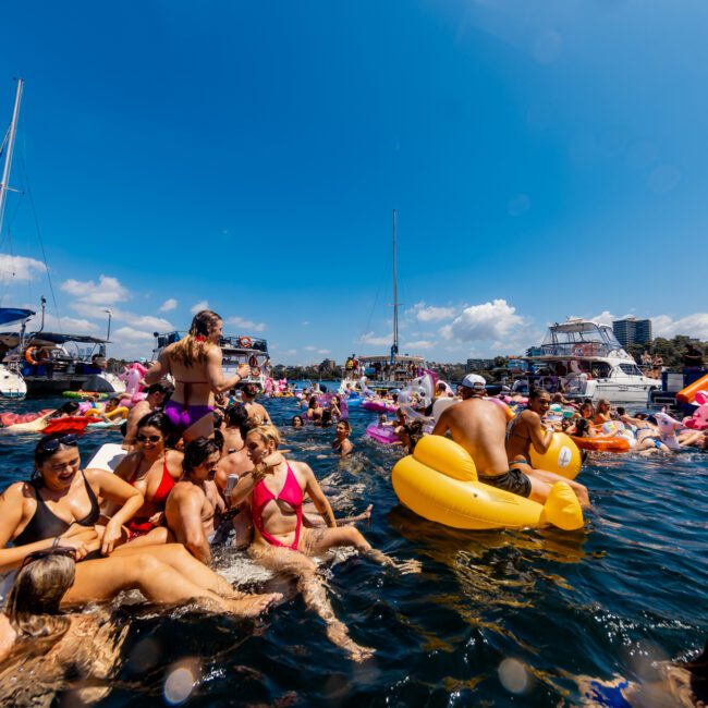 A lively scene of people enjoying a sunny day on the water, surrounded by boats and inflatables. The crowd is swimming and lounging on colorful floaties, creating a festive atmosphere. With clear skies and deep blue water, it's an ideal day for Sydney Harbour Boat Hire from The Yacht Social Club.