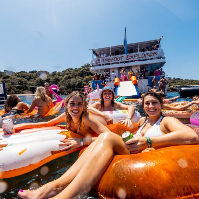 A lively group of people enjoy a sunny day on inflatable floaties in the water near a boat named "Barefoot Explorer." They're smiling and holding drinks, with lush greenery in the background. The atmosphere is festive and relaxed. A banner reads "The Yacht Social Club," perfect for Sydney Harbour Boat Hire The Yacht Social Club events.