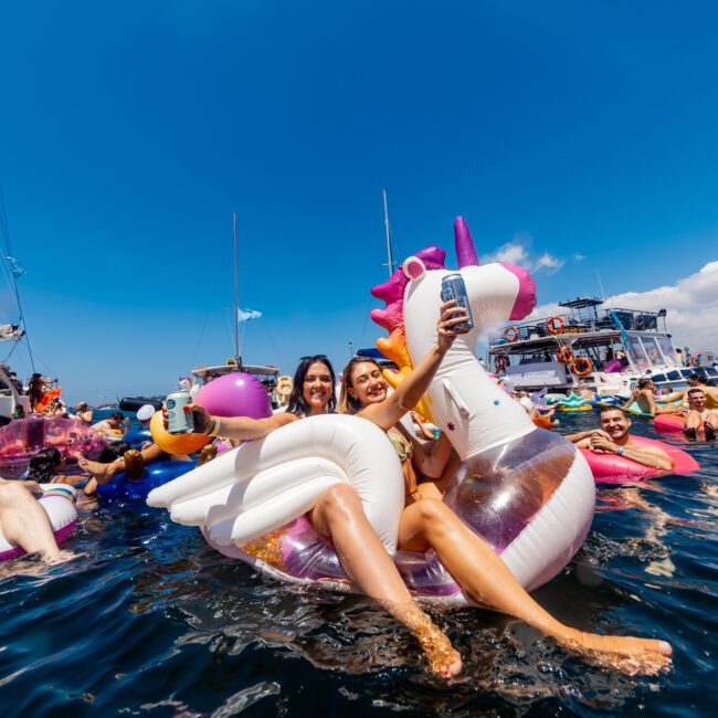 Two women sit on a unicorn pool float at The Yacht Social Club event in Sydney. They are taking selfies and holding drinks amidst a lively gathering of people on other floats in the water. Boats can be seen in the background under a clear blue sky, and the atmosphere is festive and joyful.