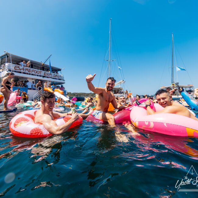 A group of people enjoying a sunny day on the water, with some in colorful inflatable floaties near a boat named "Barefoot Explorer." The Yacht Social Club and other boats in the background add to the lively, festive atmosphere.