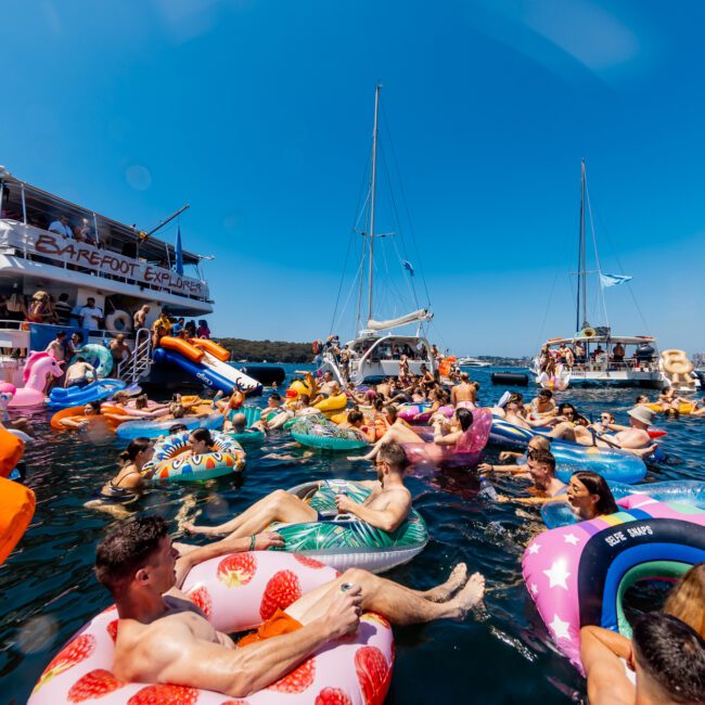 A large group of people floating on inflatable rafts of various shapes and colors in a body of water near several boats. The scene is festive, with bright sunny weather and clear blue skies. Some of the inflatables include a flamingo, donut, and unicorn; it feels like one of those vibrant Boat Parties Sydney The Yacht Social Club hosts.