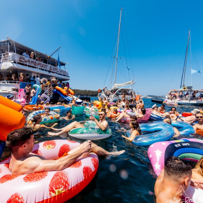 A lively group of people enjoying their time in the water on colorful inflatable floats, including tubes shaped like strawberries and pineapples, near several boats on a sunny day. The background features clear blue skies and other nearby vessels with people aboard, as part of The Yacht Social Club Event Boat Charters.