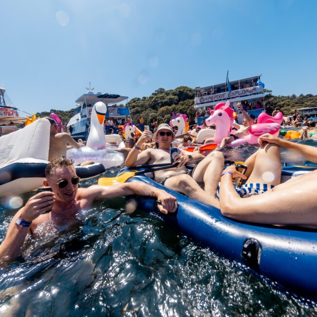 People are lounging on colorful inflatable rafts and floaties in the water, enjoying a sunny day at The Yacht Social Club Sydney Boat Hire event. Yachts are anchored nearby, and participants are wearing swimwear and sunglasses, creating a festive atmosphere. A "Yacht Social Club" sign is visible.