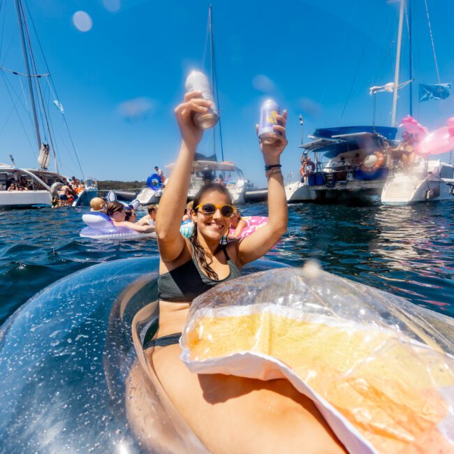 A woman in sunglasses and swimwear floats on an inflatable in the water, holding up two cans with a joyful expression. Around her, people enjoy the water with various watercraft and inflatables under a bright blue sky, as sailboats from The Yacht Social Club Sydney Boat Hire glide gracefully in the background.