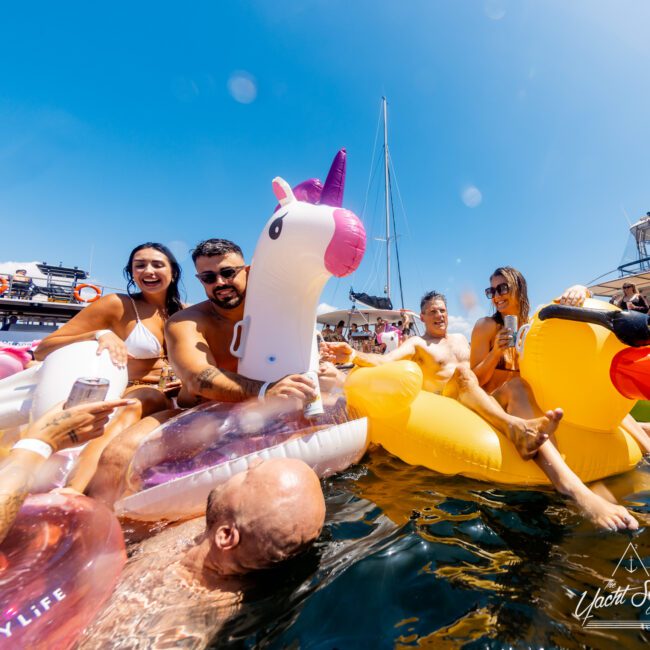 A group of people enjoying a sunny day on colorful inflatable floats, including a unicorn and a duck, in the water. They are smiling and holding drinks while surrounded by boats. The atmosphere appears fun and lively. "The Yacht Social Club Sydney Boat Hire" is visible in the corner.