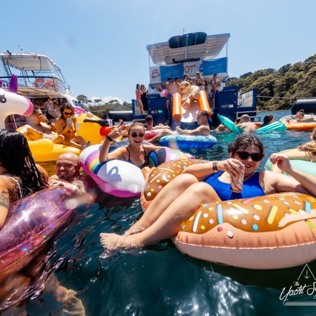 A group of people enjoy a sunny day on inflatable pool floats around yachts at The Yacht Social Club event. They relax and laugh while holding drinks. One woman in sunglasses gives a peace sign. The water is clear and blue, with trees visible in the background, creating a festive and lively atmosphere.