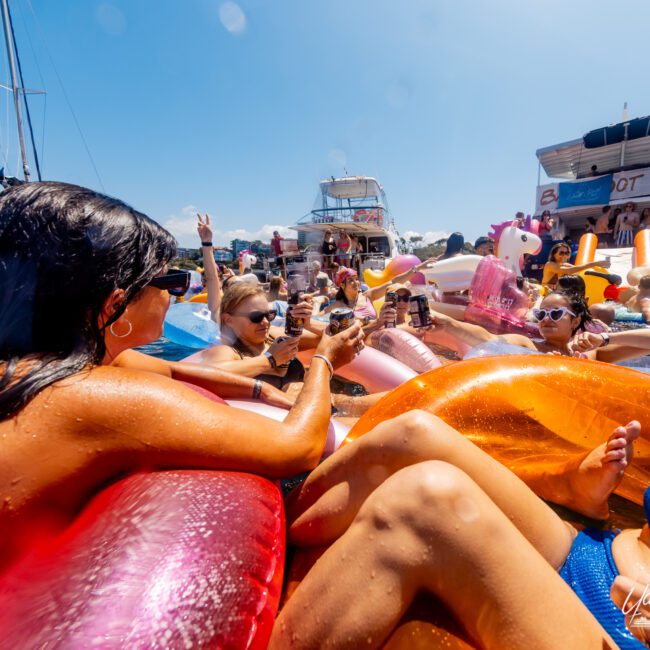 A group of people relax on colorful inflatable floats in the water, sipping drinks and enjoying the sunshine. In the background, several boats from The Yacht Social Club Sydney Boat Hire are anchored close together. Many individuals are holding up drinks and engaging in conversation, creating a festive atmosphere.