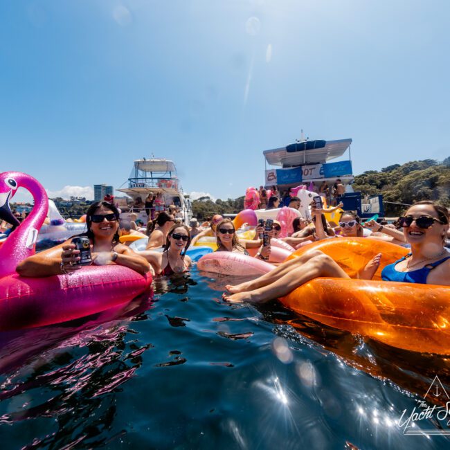 Group of people lounging on colorful inflatable rafts in a body of water, holding drinks and smiling. Boats and more inflatable rafts can be seen in the background along with green hills under a clear blue sky. A "Luxury Yacht Rentals Sydney" logo is in the bottom right corner.