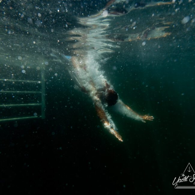 A person is swimming underwater in a dark, moody environment with their arms extended forward. Light refracts through the water's surface, creating shimmering patterns. In the bottom right corner, a logo reading "Boat Parties Sydney The Yacht Social Club" is visible.