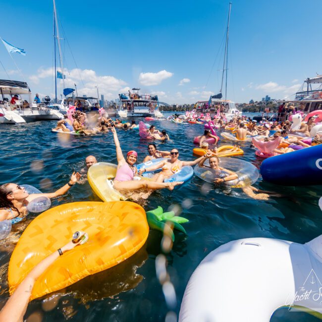 A lively gathering of people enjoying a sunny day on the water, floating on colorful inflatables shaped like pineapple slices, donuts, and flamingos. There are various boats in the background from Sydney Harbour Boat Hire The Yacht Social Club with people mingling and relaxing, creating a festive atmosphere.