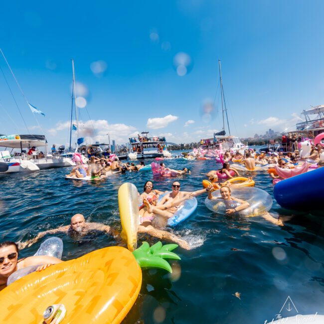 A lively scene at a lake with several people floating on colorful inflatables, including a pineapple and a donut. Boats are stationed in the background under a clear blue sky, reminiscent of a day with The Yacht Social Club Sydney Boat Hire, where everyone enjoys the sunny water.