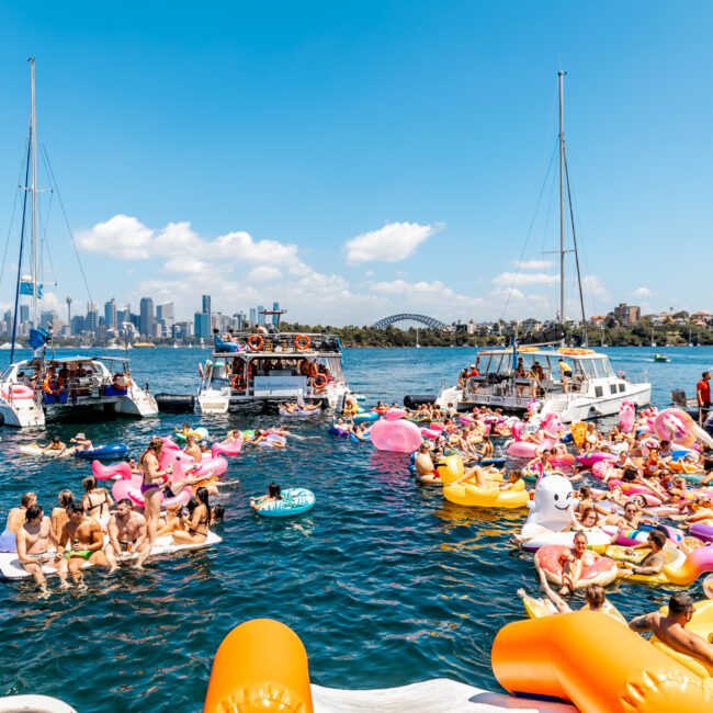 A lively scene on the water features people in swimsuits on colorful inflatable floats, surrounded by boats from The Yacht Social Club Sydney Boat Hire. The background reveals a city skyline with tall buildings beneath a clear blue sky.