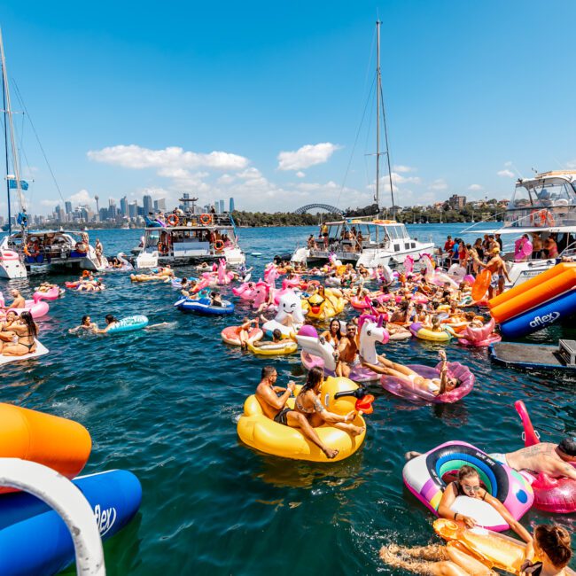 A lively scene of a yacht party on the water. Numerous people are enjoying themselves on colorful inflatable floats, including flamingos and various pool toys. Boats and yachts are in the background with a city skyline visible under a bright, clear blue sky—perfect for Luxury Yacht Rentals Sydney.