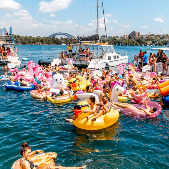 A group of people enjoying a sunny day on various colorful inflatables in calm waters near anchored boats. The background features a city skyline and a bridge under a partly cloudy sky. The scene is lively and festive, enhanced by The Yacht Social Club Event Boat Charters with many bright unicorn and donut floaties.