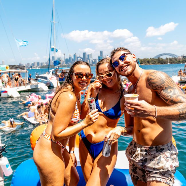 Three people in swimwear, two women and one man, smiling and enjoying drinks on a boat rented from Luxury Yacht Rentals Sydney, with a lively crowd and other boats on the water in the background. They are under a sunny, clear sky with the city skyline visible in the distance.