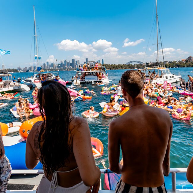 A lively boat party on a sunny day, with people on yachts and in the water on inflatables. The scene features a crowded marina, blue skies, and a city skyline in the background. Swimmers and partygoers are enjoying the festive atmosphere of The Yacht Social Club Event Boat Charters.