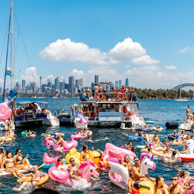 A vibrant scene of a boat party on a sunny day features numerous people on colorful inflatable rafts and unicorn floats in the water. Boats are anchored nearby, with the city skyline and a bridge visible in the background, showcasing the essence of Boat Parties Sydney The Yacht Social Club.