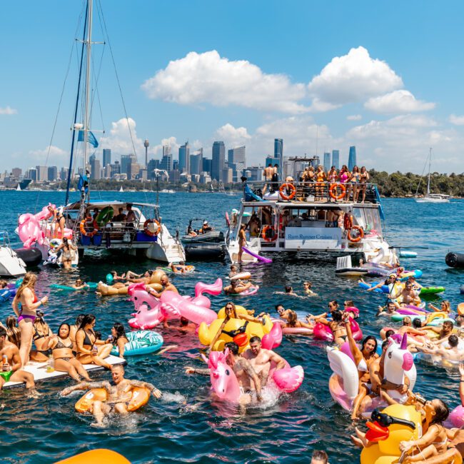 A lively scene of people partying on boats and colorful inflatable floats in the water. The backdrop features a city skyline with tall buildings against a blue sky. Hosted by Sydney Harbour Boat Hire The Yacht Social Club, the event includes swimmers and whimsical inflatables like swans and unicorns.