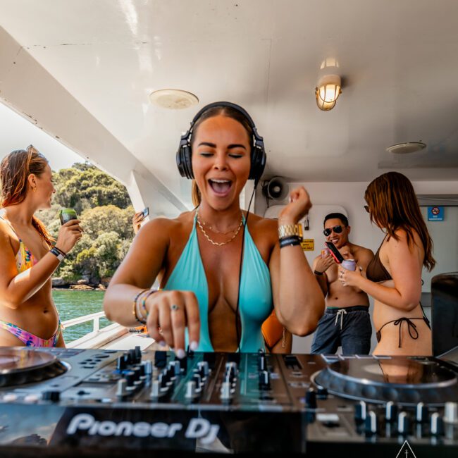 A woman in a blue bikini is DJing on a boat, wearing headphones and smiling with her eyes closed. In the background, other people in swimwear are dancing and socializing. The setting appears to be The Yacht Social Club Event Boat Charters with sunny weather and scenic views.