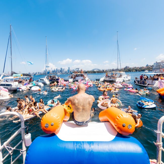 A lively and colorful gathering of people floating on inflatable rafts and swimming in a marina, surrounded by various boats. A man sits on an inflatable tied to a boat in the foreground, overlooking the festivities under a bright, clear sky, courtesy of The Yacht Social Club Sydney Boat Hire.